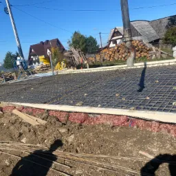 Przygotowanie zbrojenia i szalunku pod wylewkę fundamentów domu jednorodzinnego, widoczna pompa do betonu, teren budowy z ziemią i elementami drewnianymi.