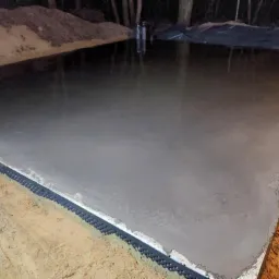 Świeżo wylana, szara wylewka betonowa z czarną listwą dylatacyjną przy krawędzi, na tle ciemnego otoczenia i drzew. Widok z góry, lekki połysk betonu.