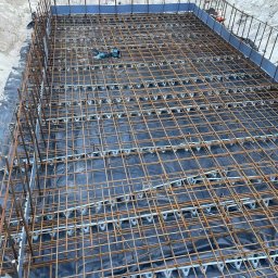 Budownictwo Tomczyk Przemysław - Zbrojenie fundamentu z użyciem dystansów i folii, widoczne narzędzie do cięcia. Krawędzie zabezpieczone szalunkiem, grunt wokół wykopu.