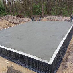 Budownictwo Tomczyk Przemysław - Świeżo wylana, gładka płyta fundamentowa z hydroizolacją, widoczna na tle piaszczystego gruntu i drzew. Pionowe rury wystają z płyty.