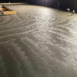 Budownictwo Tomczyk Przemysław - Świeżo wylana, mokra wylewka betonowa oświetlona lampą budowlaną, z widocznym szalunkiem i narzędziami. Charakterystyczna faktura powierzchni betonu.