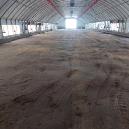 Budownictwo Tomczyk Przemysław - Przygotowanie podłoża w hali z metalową konstrukcją i oświetleniem, widoczne ślady opon. W tle jasne światło wpadające przez otwór.