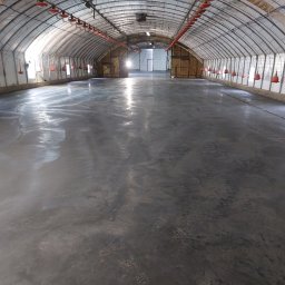 Budownictwo Tomczyk Przemysław - Nowa, gładka posadzka przemysłowa w hali z łukowatym dachem. Widoczne czerwone karmniki pod sufitem i wpadające światło z zewnątrz. Świeża, szara wylewka.