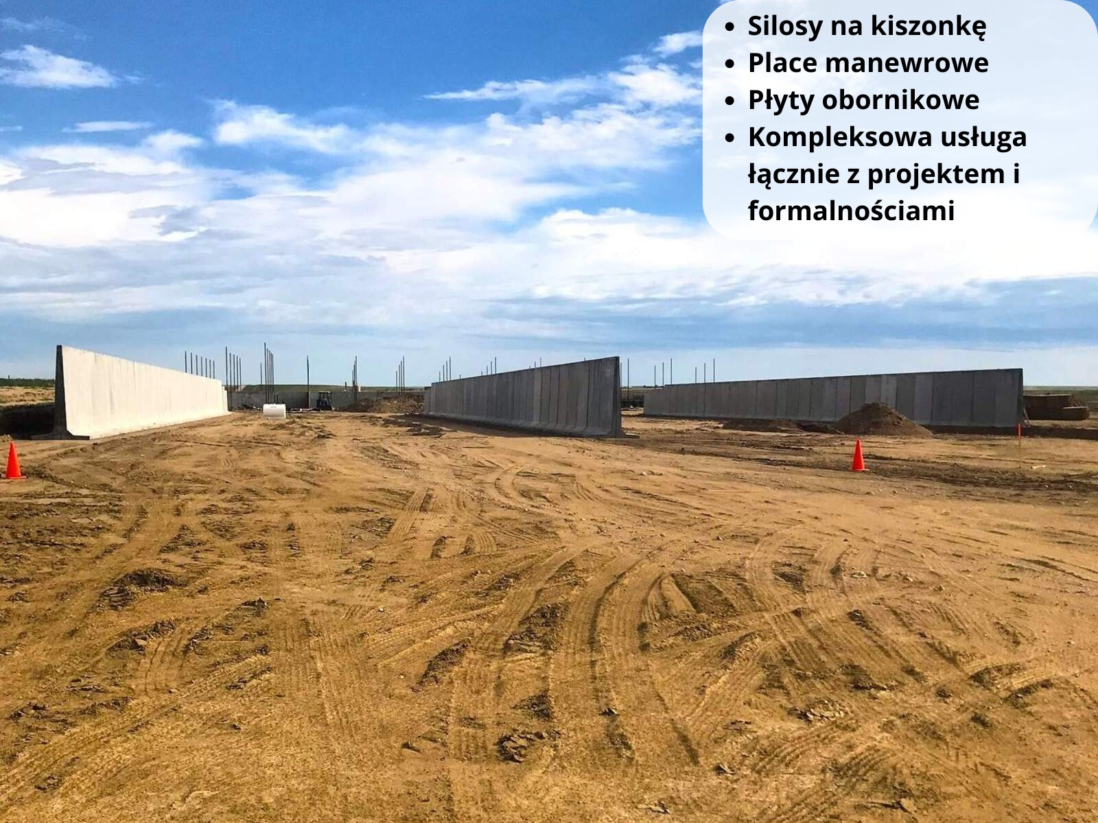Betonowe ściany oporowe w trakcie budowy na placu z widocznymi śladami opon, pomarańczowe pachołki ostrzegawcze, błękitne niebo z chmurami w tle, teren budowy.