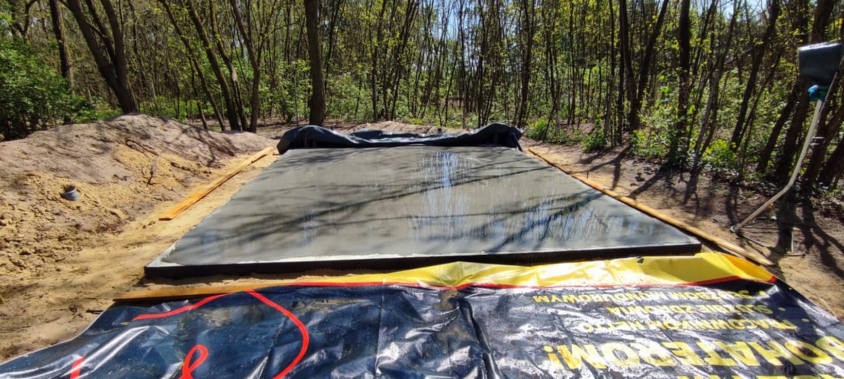 Świeżo wylana, mokra wylewka betonowa otoczona drewnianym szalunkiem na tle leśnego krajobrazu. Widoczna folia ochronna i ubity grunt.