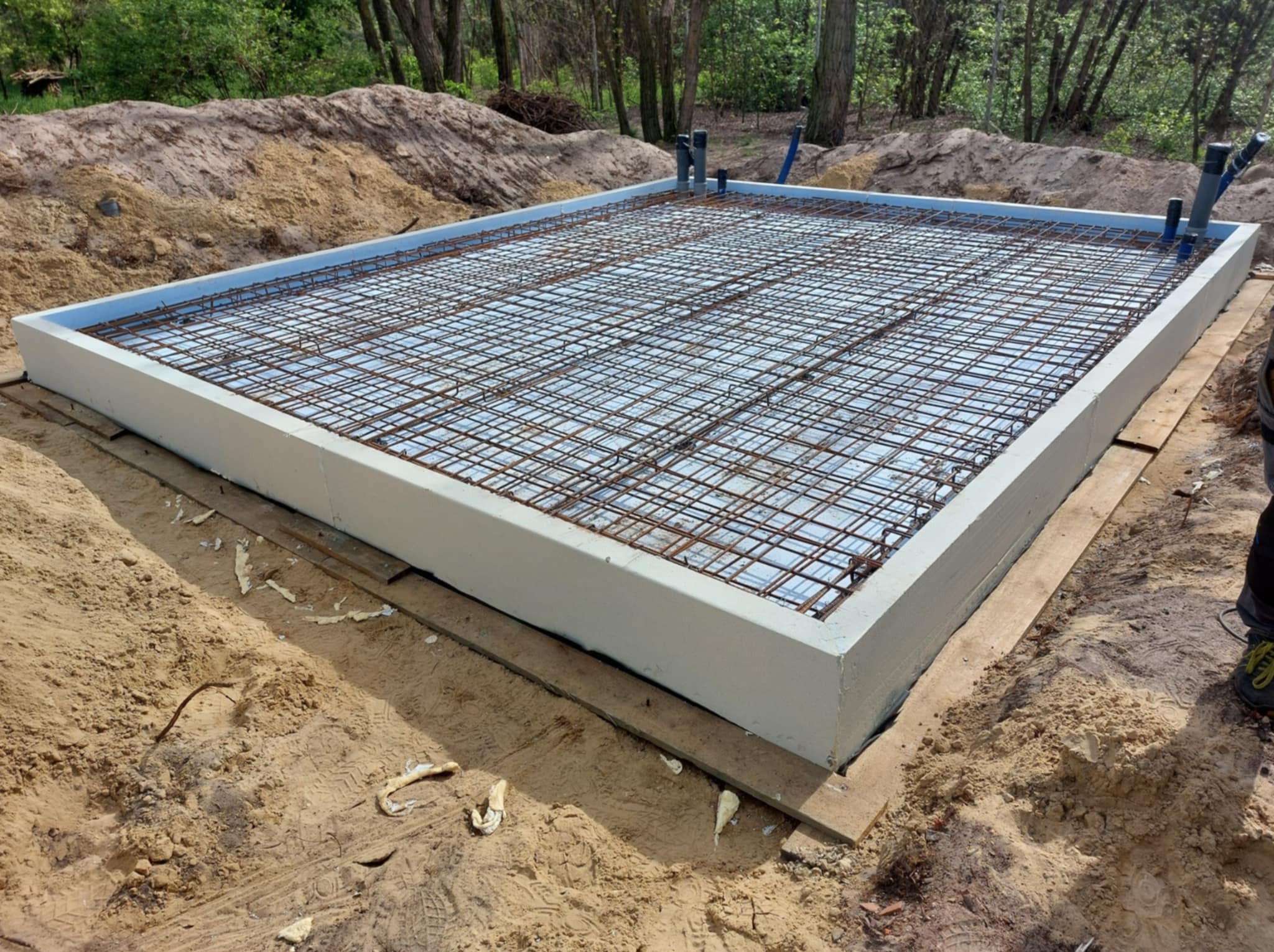 Przygotowany fundament z izolacją i zbrojeniem stalowym, widoczne rury instalacyjne, całość otoczona piaskiem. Gotowy etap prac fundamentowych.