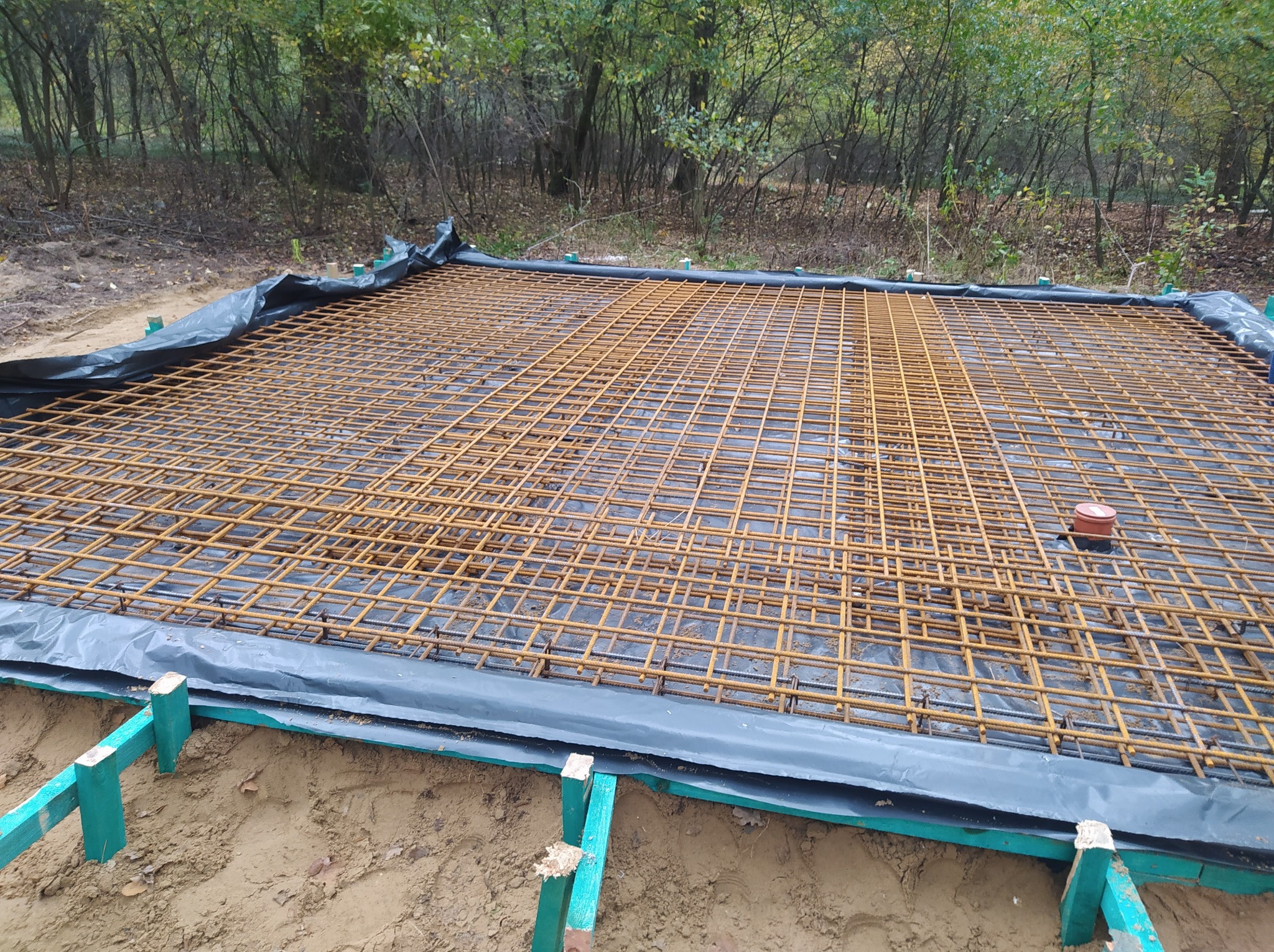 Zbrojenie pod płytę fundamentową na czarnej folii, zielone drewniane szalunki, w tle drzewa. Widok z góry, ukośny kadr.