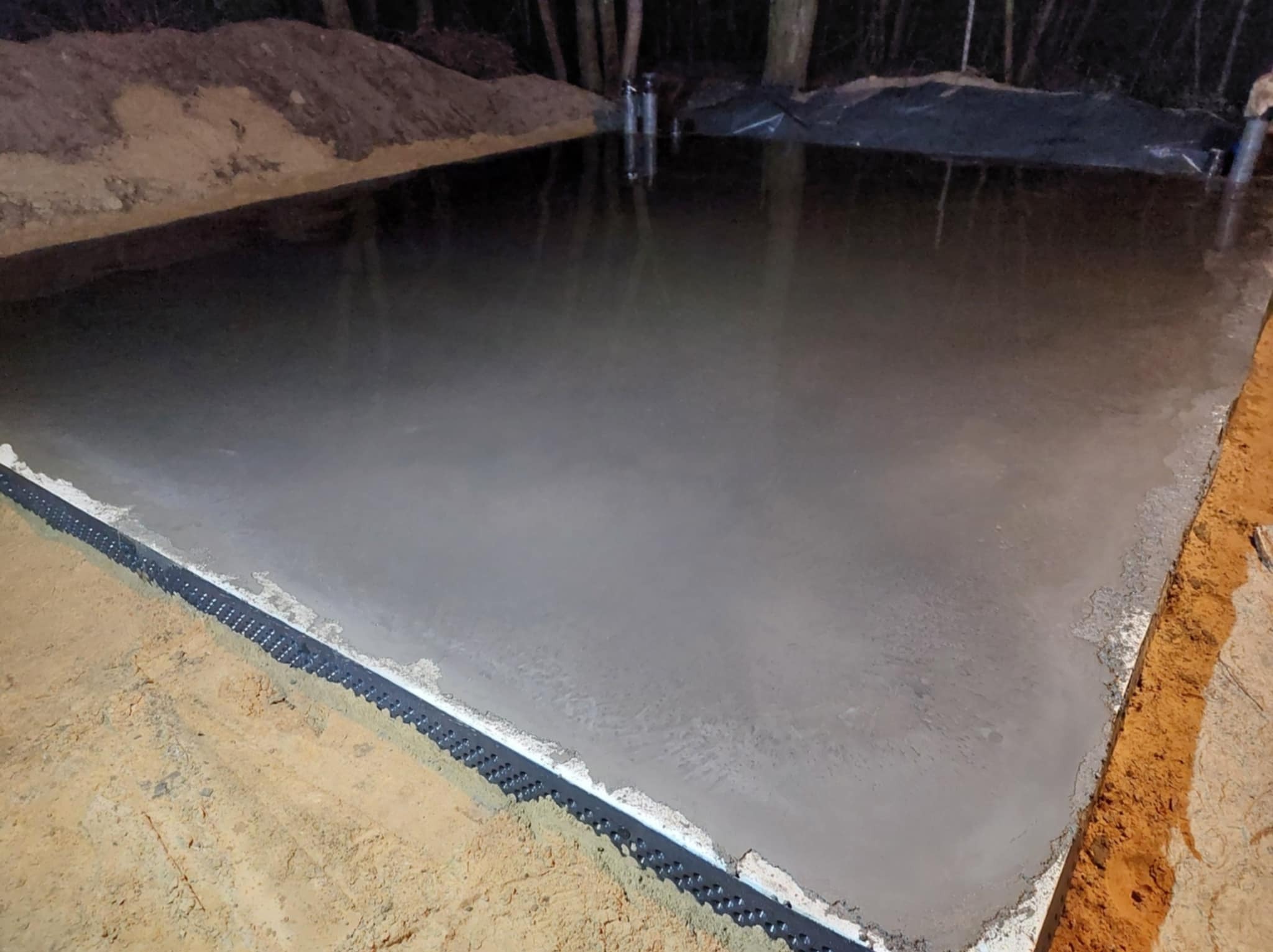 Świeżo wylana, szara wylewka betonowa z czarną listwą dylatacyjną przy krawędzi, na tle ciemnego otoczenia i drzew. Widok z góry, lekki połysk betonu.