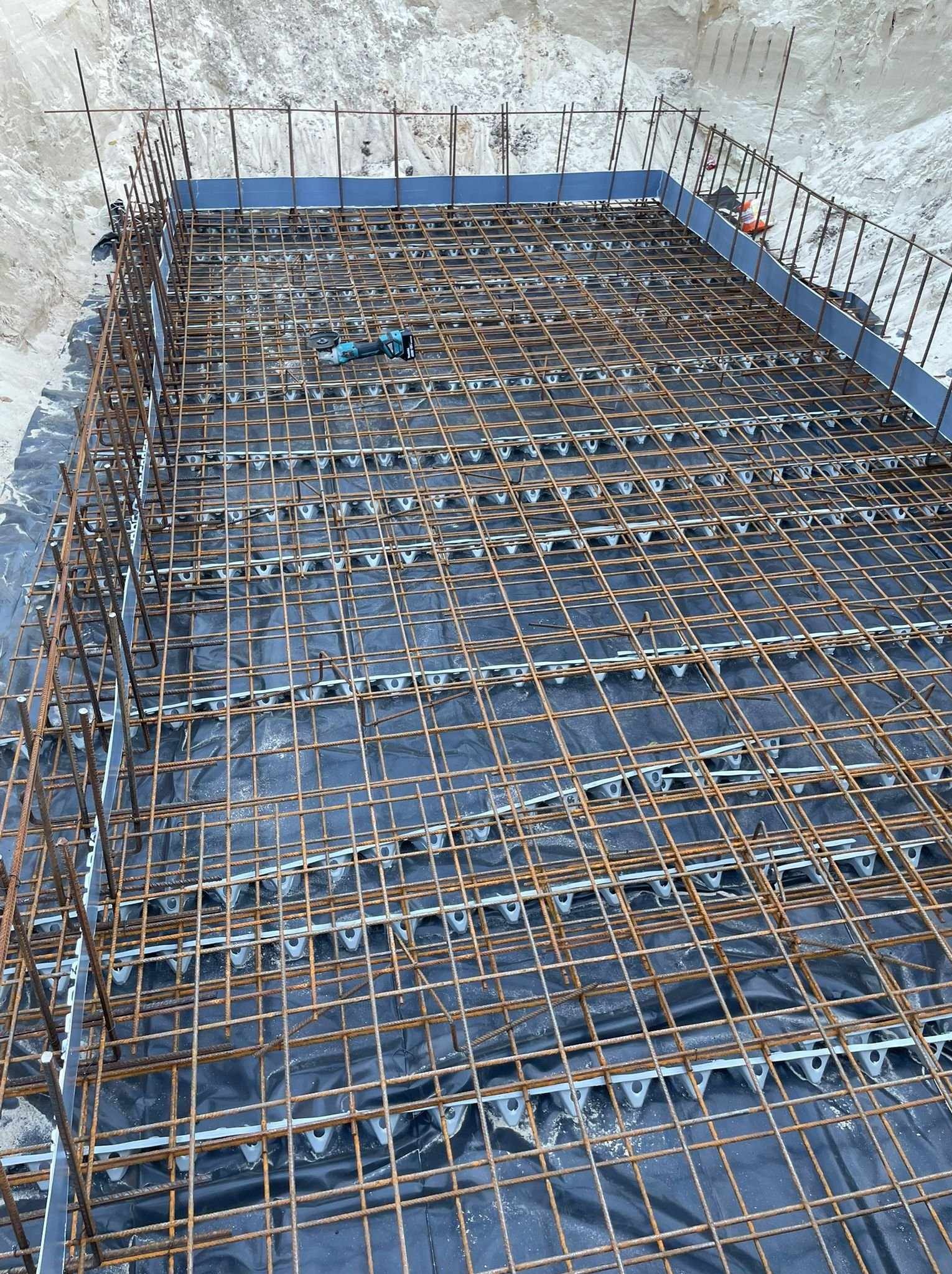 Zbrojenie fundamentu z użyciem dystansów i folii, widoczne narzędzie do cięcia. Krawędzie zabezpieczone szalunkiem, grunt wokół wykopu.