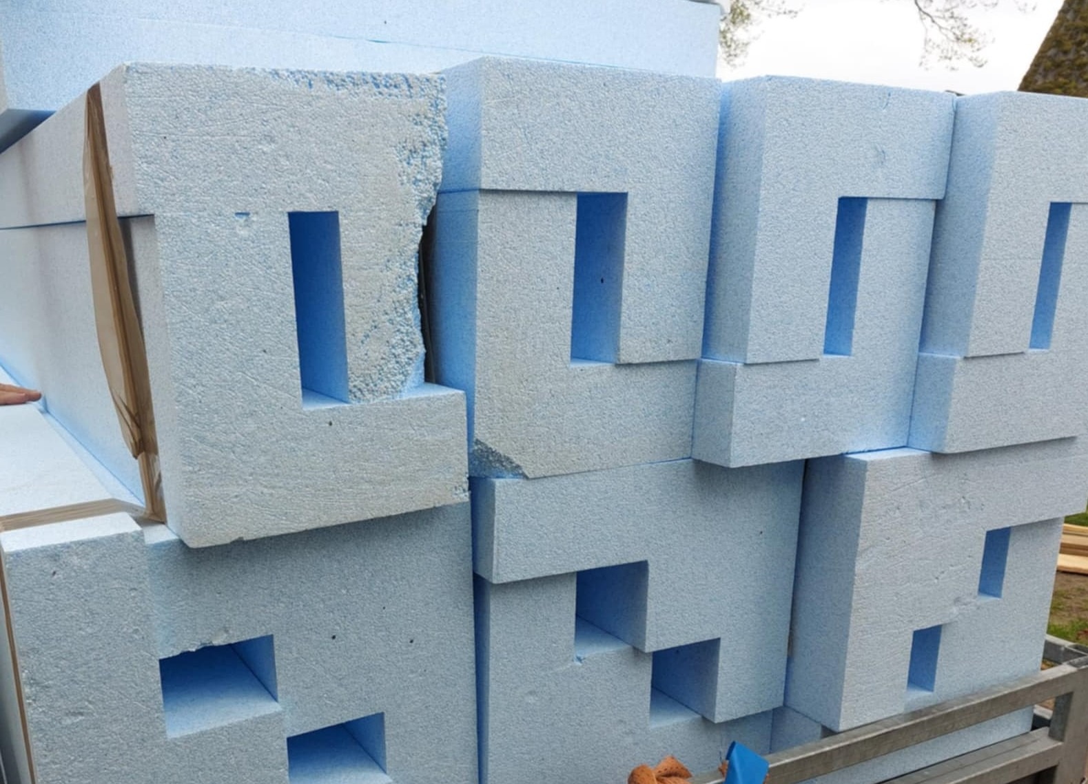 Styropianowe bloczki z wycięciami, ułożone w stos. Elementy izolacyjne w kolorze jasnoniebieskim, widoczne uszkodzenia na jednym z bloczków. Detal materiałów budowlanych.