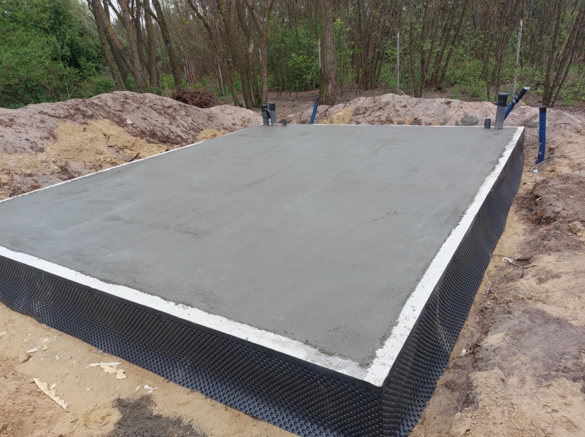 Świeżo wylana, gładka płyta fundamentowa z hydroizolacją, widoczna na tle piaszczystego gruntu i drzew. Pionowe rury wystają z płyty.