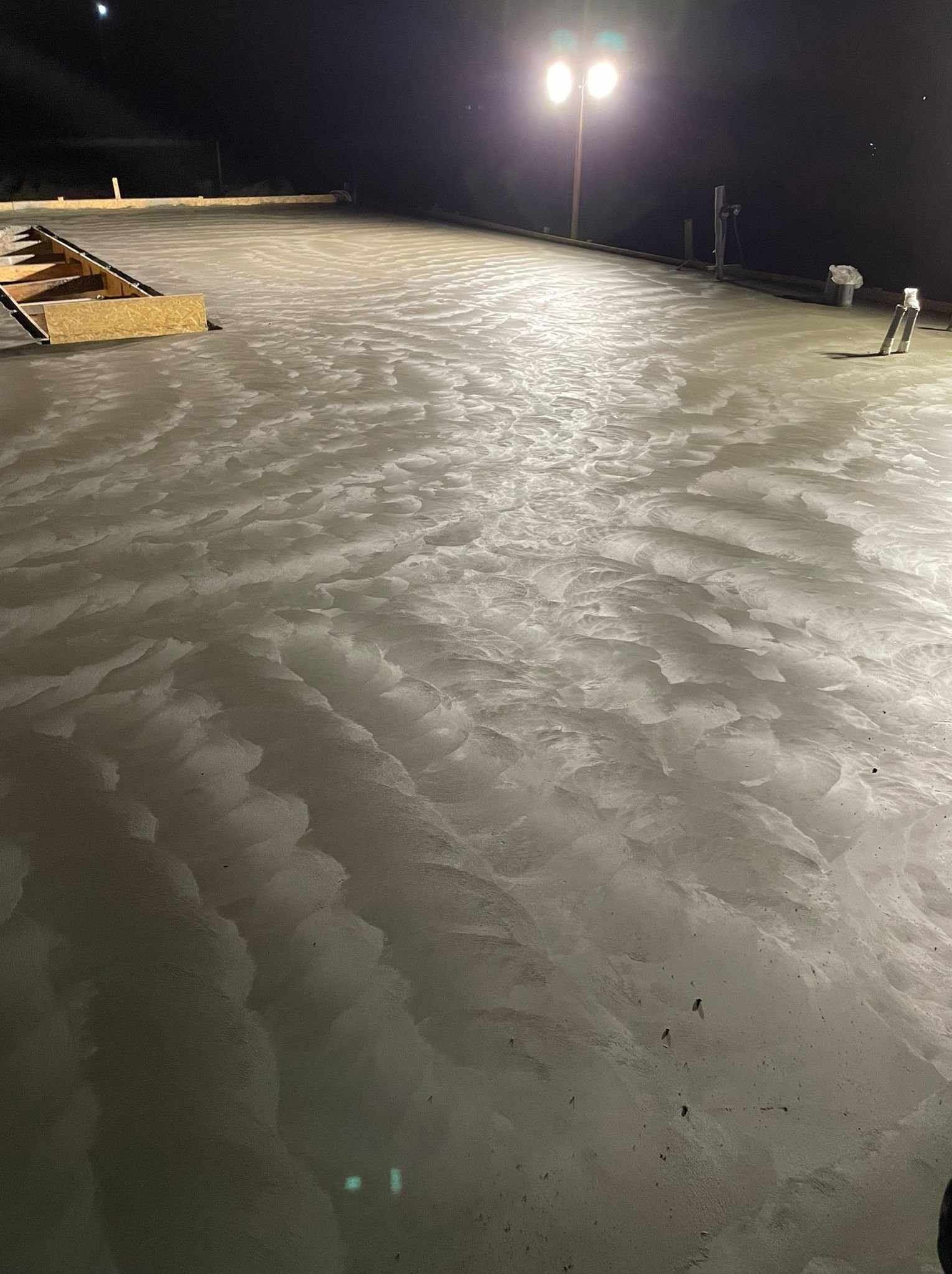 Świeżo wylana, mokra wylewka betonowa oświetlona lampą budowlaną, z widocznym szalunkiem i narzędziami. Charakterystyczna faktura powierzchni betonu.