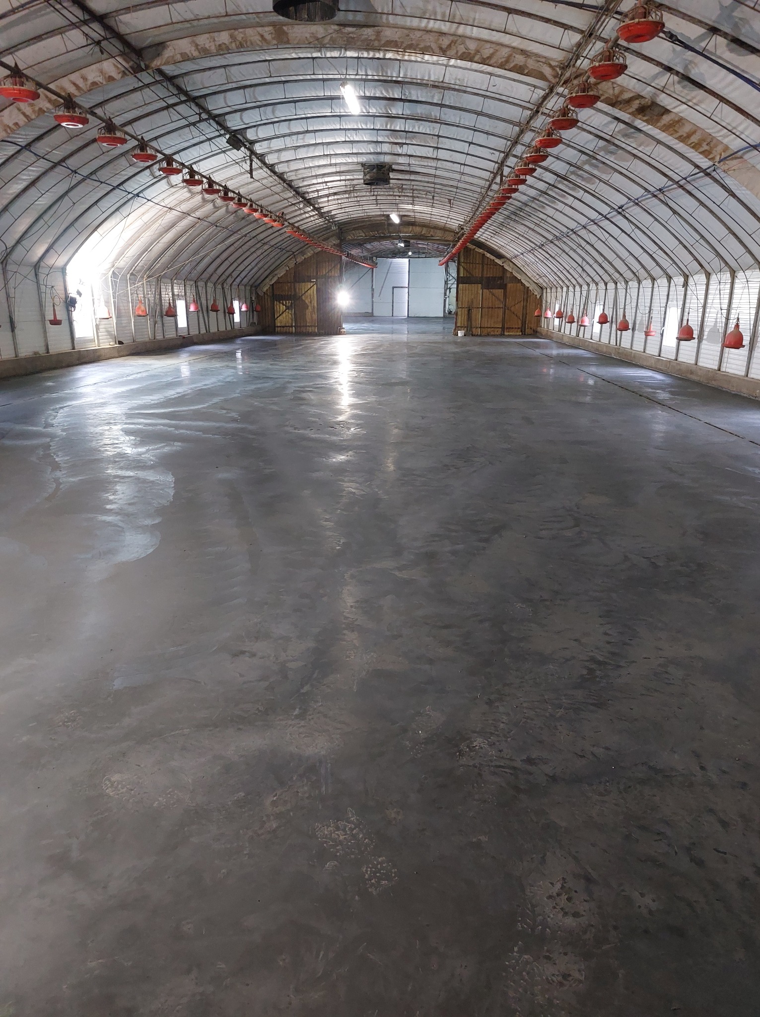 Nowa, gładka posadzka przemysłowa w hali z łukowatym dachem. Widoczne czerwone karmniki pod sufitem i wpadające światło z zewnątrz. Świeża, szara wylewka.