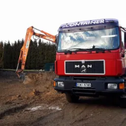 Czerwona ciężarówka MAN z naczepą wywrotką i pomarańczowa koparka ATLAS na placu budowy, obok wykopu pod fundamenty domu, w tle rząd drzew.