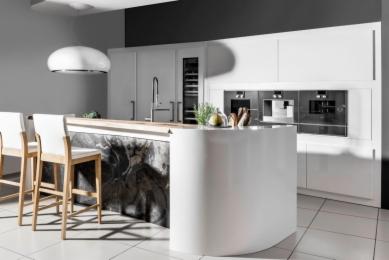 Nowoczesna, minimalistyczna kuchnia z białymi, błyszczącymi frontami, wyspą z zaokrąglonymi krawędziami i blatem z jasnego drewna, podkreślona marmurkowym wykończeniem wyspy i białymi krzesłami...