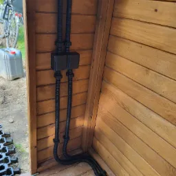 Dwie czarne rury karbowane prowadzone wewnątrz drewnianej konstrukcji, zabezpieczone czarnymi puszkami i uchwytami, widok z dołu na instalację w trakcie montażu.