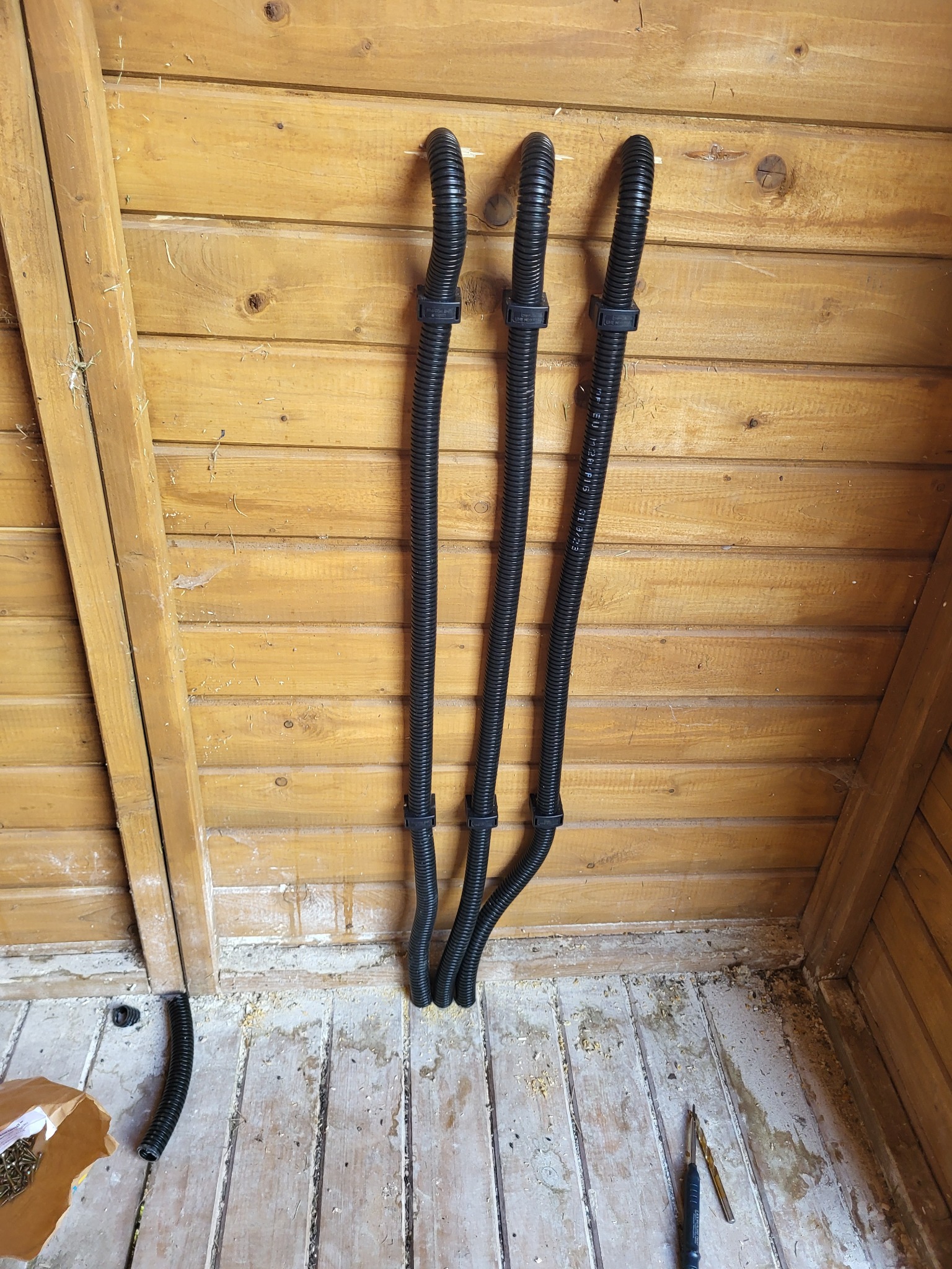 Trzy czarne, karbowane rury ochronne przymocowane do drewnianej ściany za pomocą uchwytów, widoczne w rogu pomieszczenia, w tle drewniana ściana, na podłodze wiertła i rozsypane wkręty.