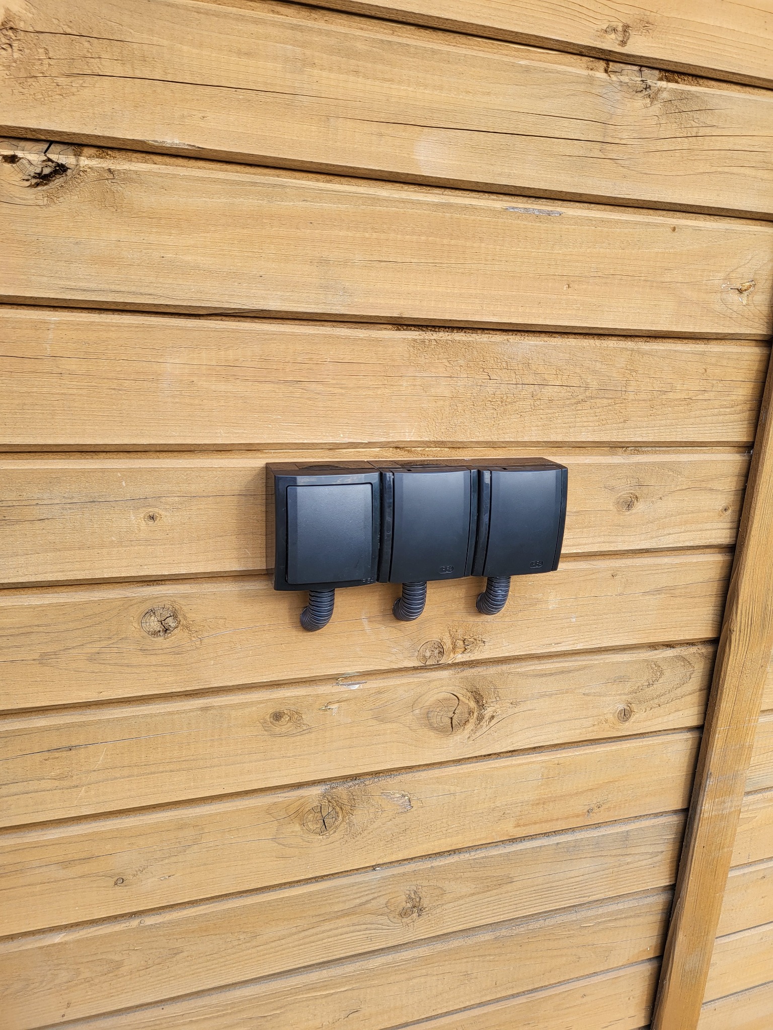 Trzy czarne puszki elektryczne natynkowe z peszlami zamontowane na drewnianej elewacji budynku.