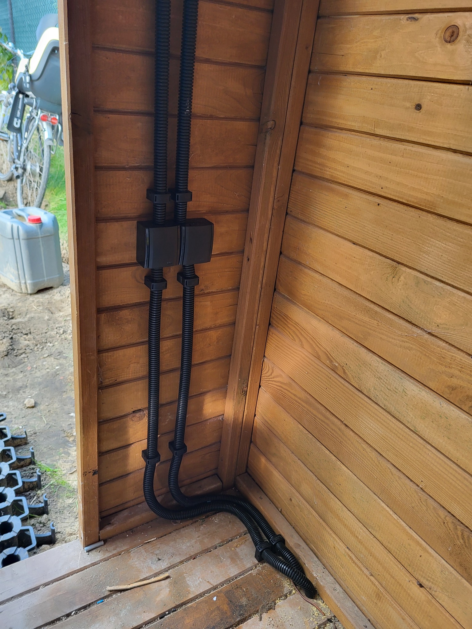 Dwie czarne rury karbowane prowadzone wewnątrz drewnianej konstrukcji, zabezpieczone czarnymi puszkami i uchwytami, widok z dołu na instalację w trakcie montażu.