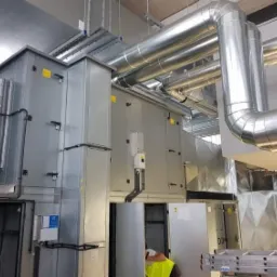 Rozbudowany system wentylacyjny z metalowymi rurami i centralą wentylacyjną w pomieszczeniu przemysłowym, widoczna drabina i fragment osoby w kamizelce odblaskowej.