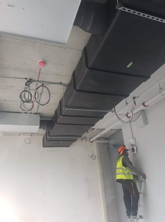 Czarny, prostokątny kanał wentylacyjny zamontowany pod betonowym sufitem w surowym wnętrzu budynku. Pracownik w kasku i kamizelce na drabinie.