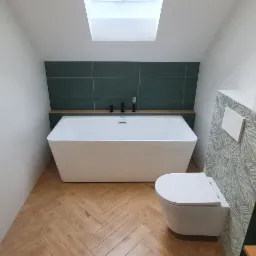 Nowoczesna łazienka na poddaszu z białą wanną wolnostojącą, toaletą podwieszaną, zielonymi płytkami i drewnianą podłogą w jodełkę, oświetlona światłem z okna dachowego.