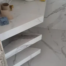 Nowoczesna łazienka z marmurowymi płytkami, białą umywalką nablatową i złotą armaturą, widoczne niedokończone prace przy obudowie stelaża.