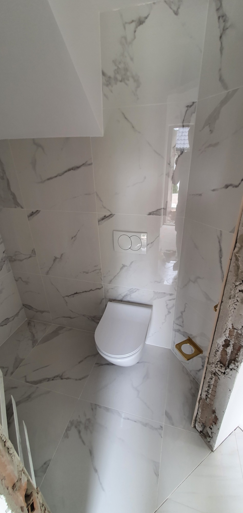 Nowoczesna toaleta wisząca z podtynkową spłuczką w łazience wykończonej białymi płytkami imitującymi marmur, widoczny fragment niezakończonego wykończenia przy drzwiach.