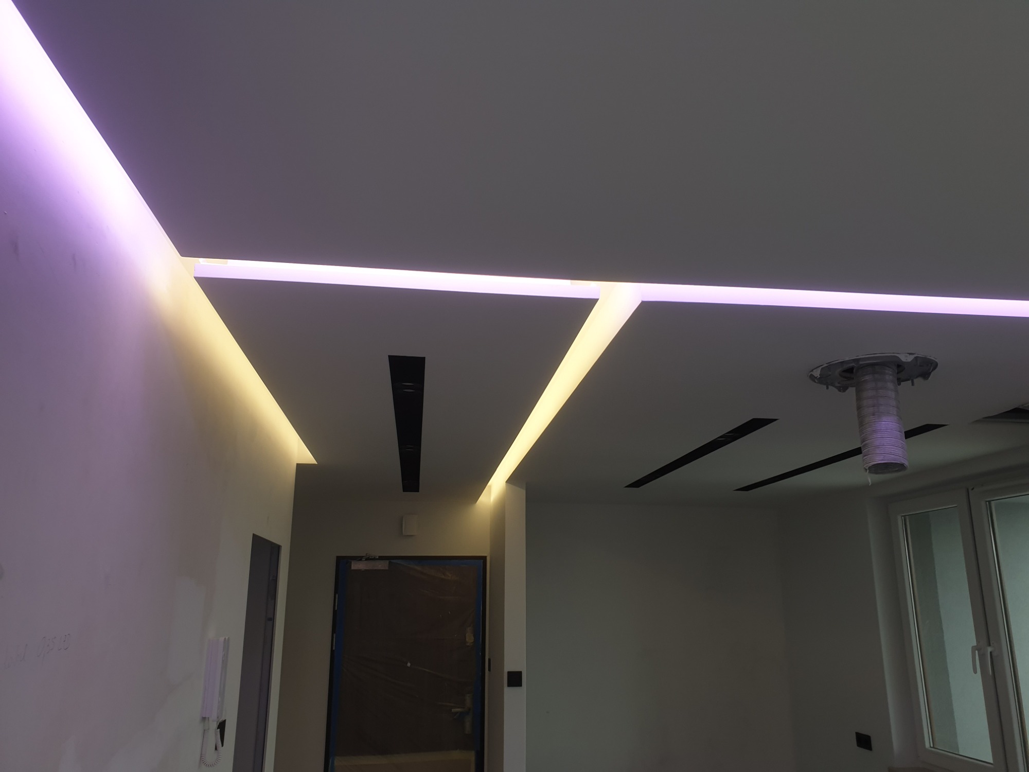 Nowoczesny sufit podwieszany z oświetleniem LED w kolorze fioletowym i żółtym, widoczne elementy instalacji wentylacyjnej i czarne listwy oświetleniowe.