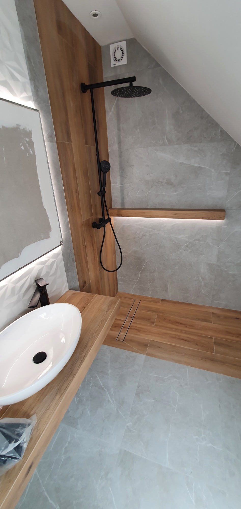 Nowoczesna łazienka w odcieniach szarości i drewna, z czarną armaturą, owalną umywalką nablatową i odpływem liniowym w strefie prysznica, oświetlenie LED pod półką.