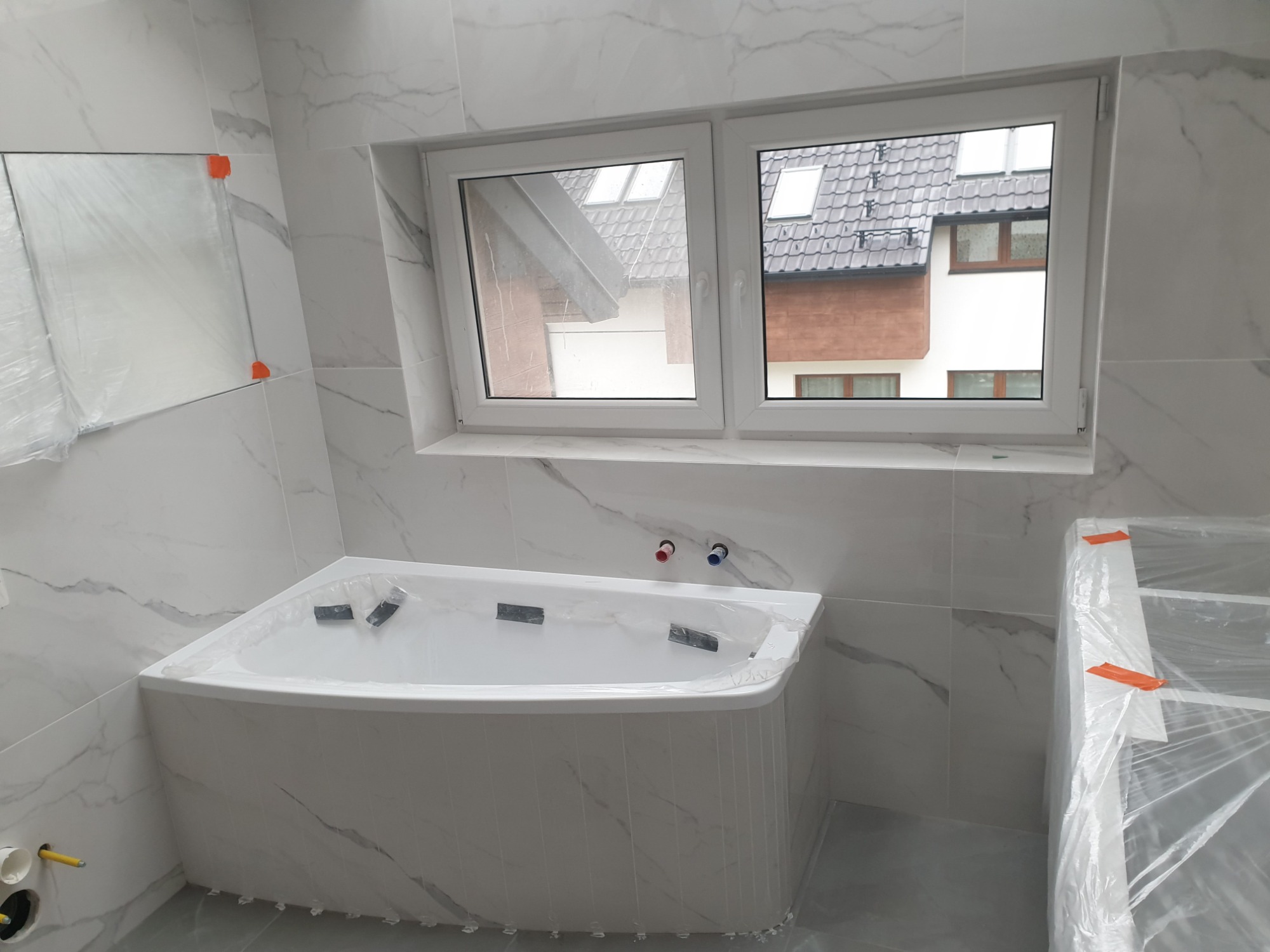 Nowa, narożna wanna z hydromasażem, zabezpieczona folią ochronną, w łazience wykończonej białymi płytkami imitującymi marmur, widok na okno z widokiem na sąsiedni dom.