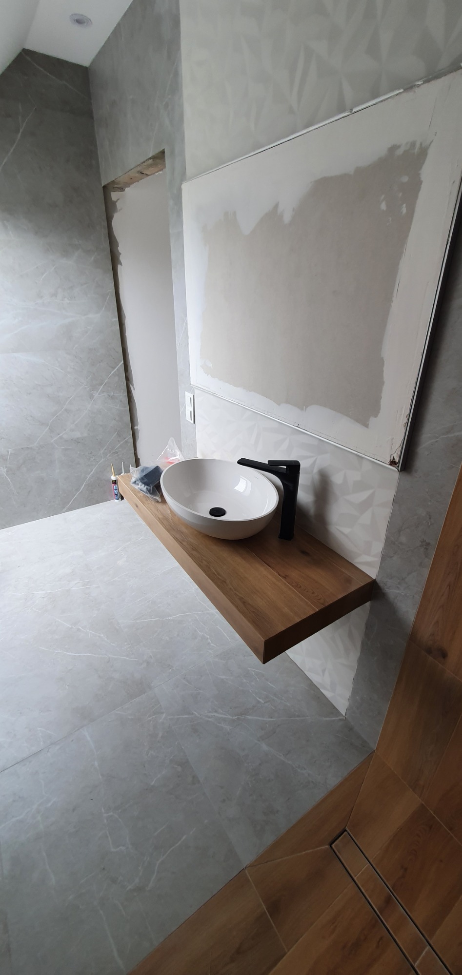 Łazienka w trakcie remontu: biała umywalka nablatowa z czarną armaturą na drewnianym blacie, szare płytki imitujące marmur na ścianach i podłodze, widoczne materiały budowlane i niedokończone...