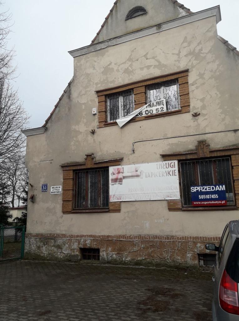 Fasada starego budynku z banerami reklamującymi usługi tapicerskie i sprzedaż nieruchomości, widoczny fragment zaparkowanego samochodu.