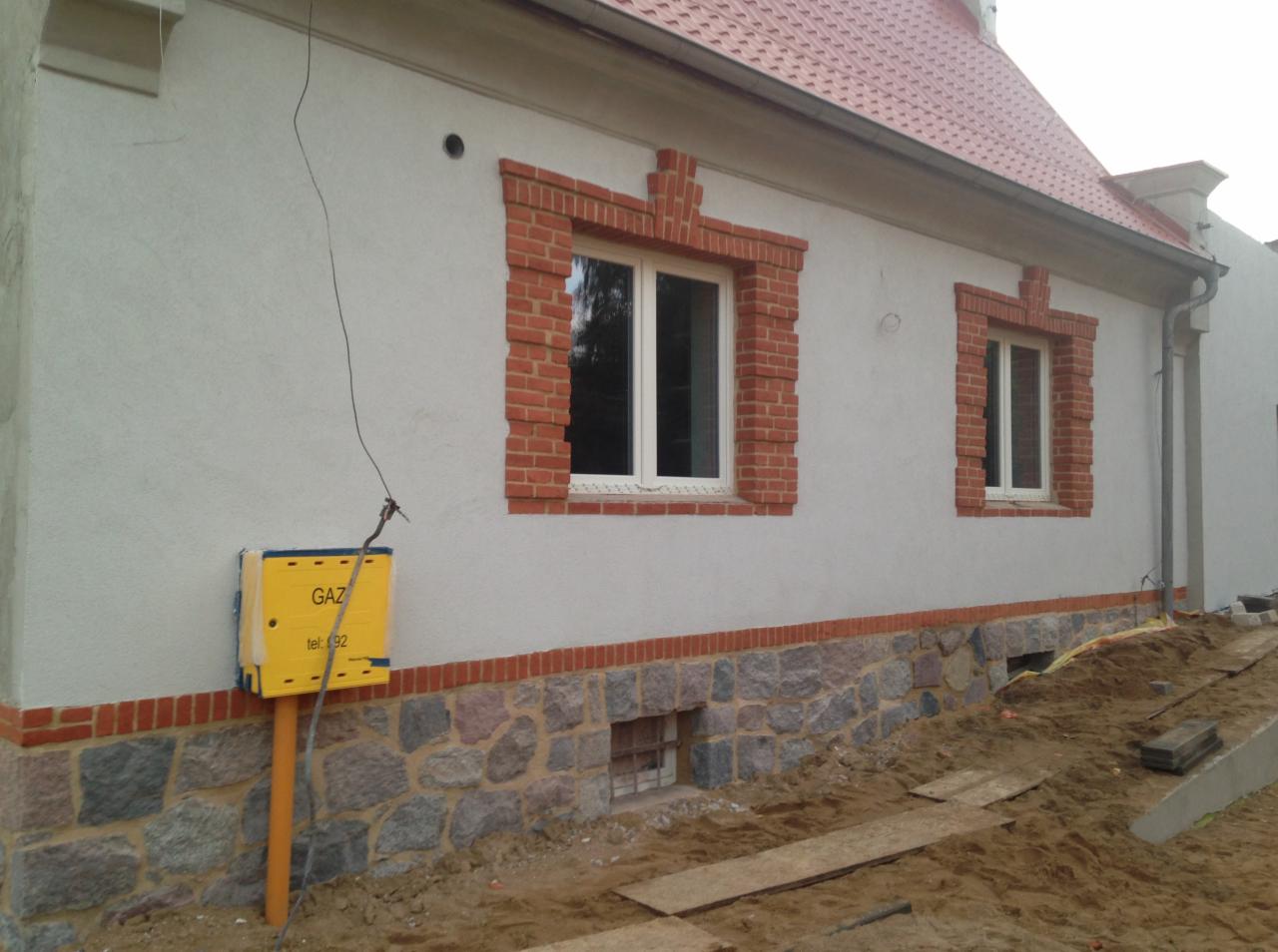 Elewacja domu z oknami obramowanymi czerwoną cegłą, cokół z kamienia, żółta skrzynka gazowa na ścianie, teren wokół domu w trakcie prac ziemnych.