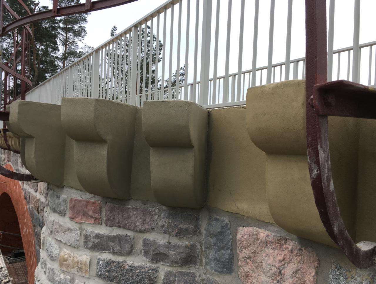 Kamienny mur z odnowionymi wspornikami pod balustradą, widoczny fragment metalowej konstrukcji schodów.