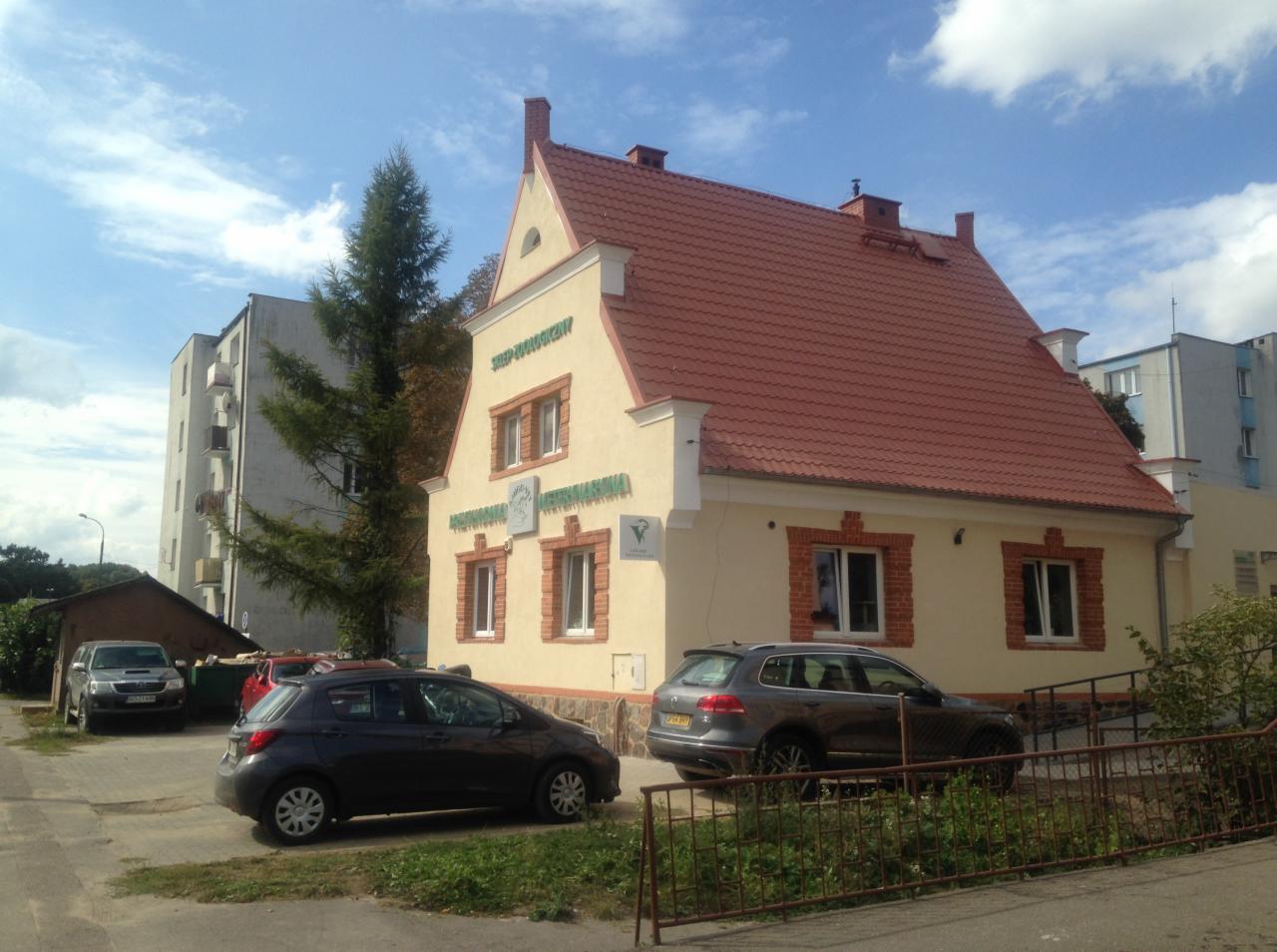 Budynek z żółtą elewacją i czerwoną dachówką, stylizowany na architekturę historyczną, z napisem 'Sklep Zoologiczny' nad oknami, zaparkowane samochody na pierwszym planie.