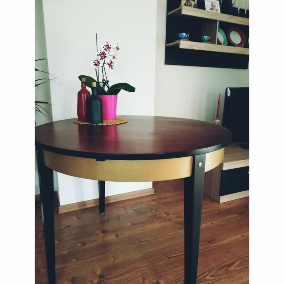 Okrągły stół z ciemnego drewna z czarnymi nogami, na blacie dwie butelki z korkami, różowa orchidea w doniczce, w tle drewniana podłoga i półki na ścianie.