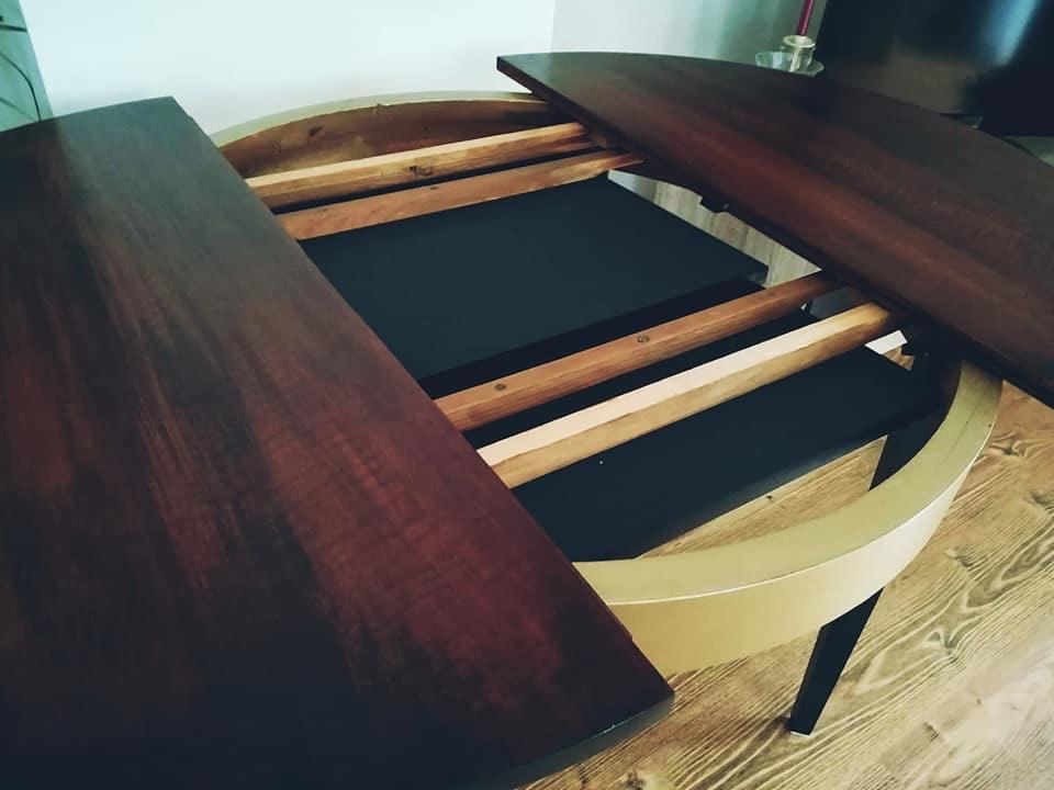 Rozłożony okrągły stół z ciemnego drewna, widoczne drewniane prowadnice i czarny blat pod spodem, złoty pierścień okalający blat, czarne nogi, parkiet podłogowy.