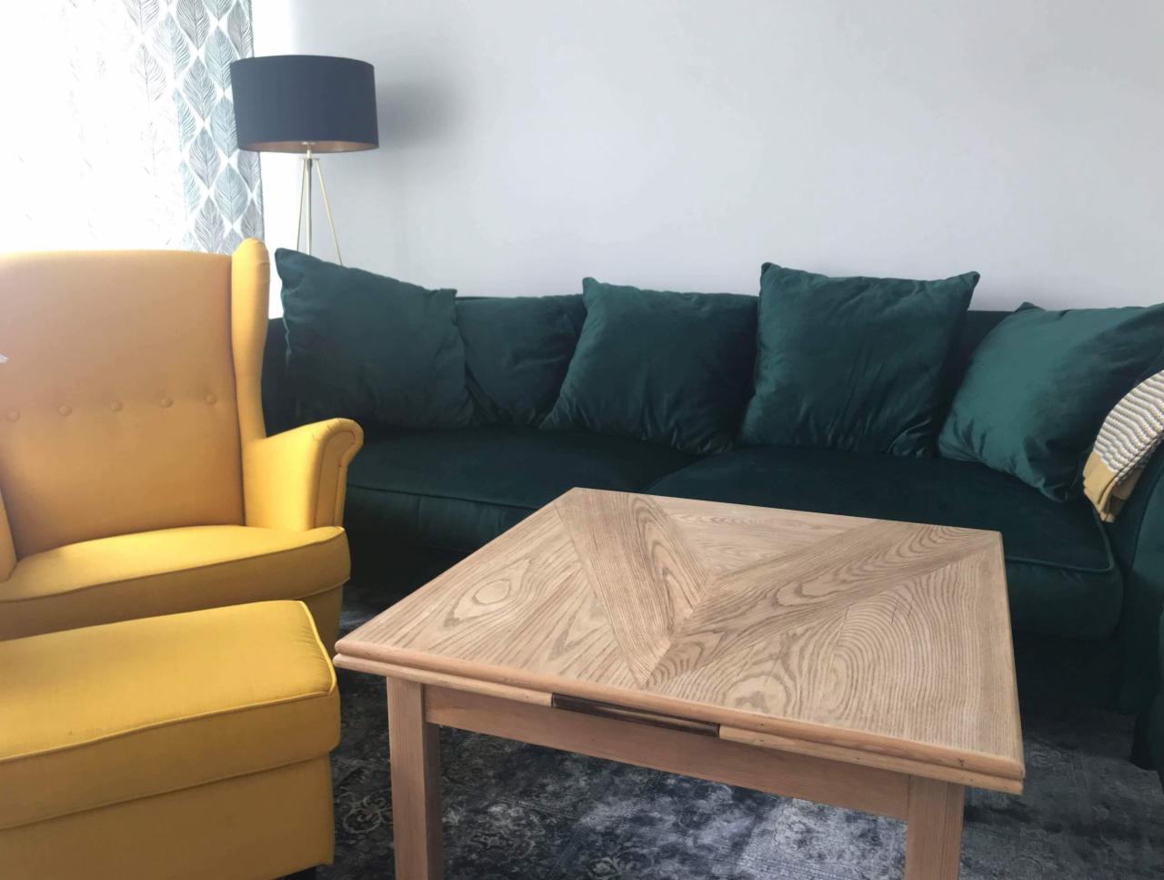 Aranżacja wnętrza w Olsztynie: żółty fotel z podnóżkiem, ciemnozielona sofa z poduszkami i drewniany stolik kawowy na wzorzystym dywanie.