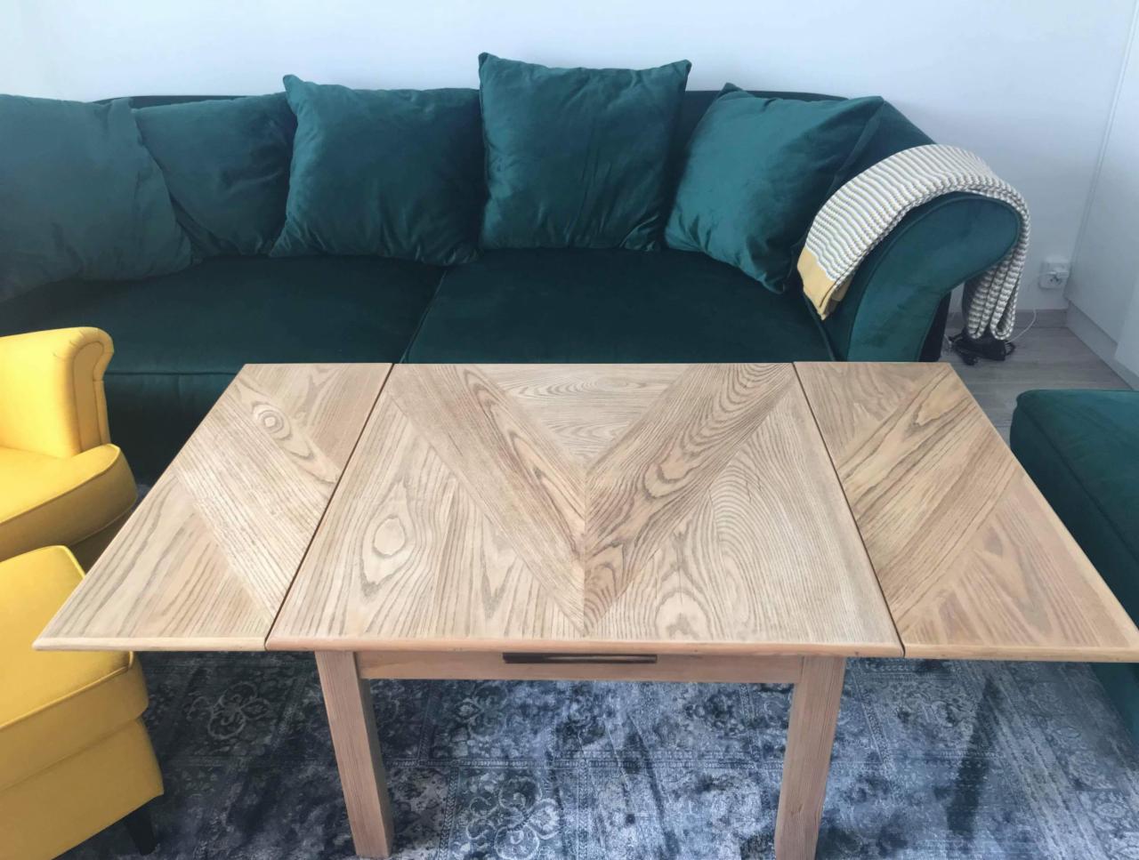 Rozkładany stół z jasnego drewna z geometrycznym wzorem blatu, na tle ciemnozielonej sofy i żółtego fotela, na wzorzystym dywanie.