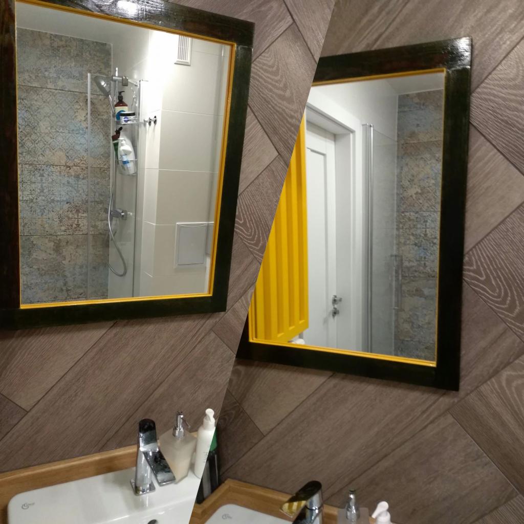Dwa lustra w ciemnych ramach z żółtymi akcentami zawieszone na ścianie z drewnopodobnych paneli ułożonych w jodełkę, odbijające kabinę prysznicową i żółte drzwi.