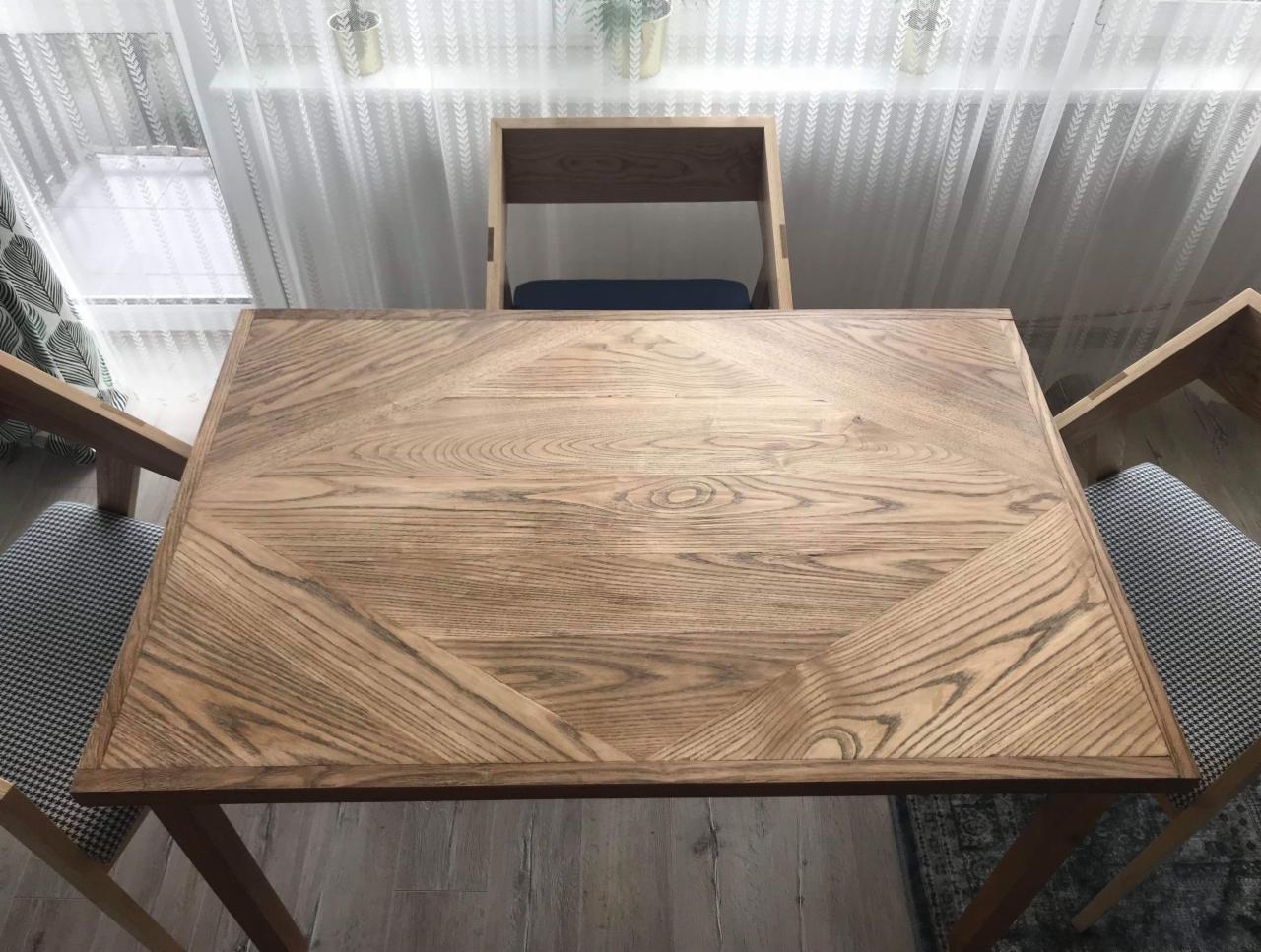 Stół z blatem z jasnego drewna o geometrycznym wzorze, otoczony krzesłami z tapicerką w pepitkę, ustawiony na tle okna z delikatną firanką.