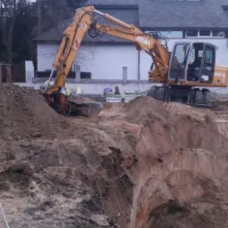 Żółta koparka Atlas 1404 M wykonuje wykop w ziemi, widoczne warstwy gleby i tyczki geodezyjne, w tle budynek mieszkalny z szarym dachem.