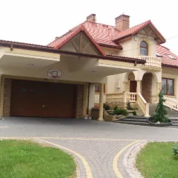 Dwupiętrowa rezydencja z garażem, balkonem i podjazdem z kostki brukowej; widoczny kosz do koszykówki nad bramą garażową.