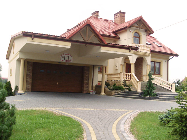 Dwupiętrowa rezydencja z garażem, balkonem i podjazdem z kostki brukowej; widoczny kosz do koszykówki nad bramą garażową.