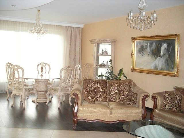 Eleganckie wnętrze salonu z okrągłym stołem i krzesłami w stylu Ludwika, kryształowe żyrandole, sofa z wzorzystymi poduszkami oraz obraz z motywem koni w złotej ramie.