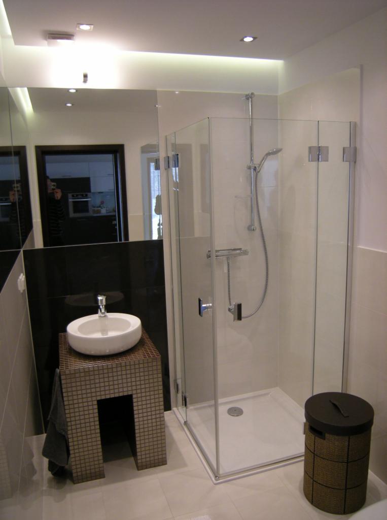 Nowoczesna łazienka z kabiną prysznicową ze szkła, umywalką nablatową na mozaikowym postumencie i dużym lustrem, w którym odbija się fragment kuchni.