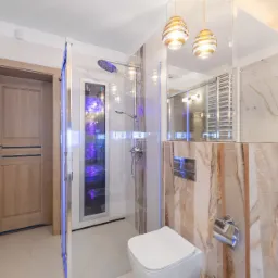 Nowoczesna łazienka w Borówcu z kabiną prysznicową podświetloną na fioletowo, marmurowe ściany, wisząca toaleta i dekoracyjne lampy wiszące.