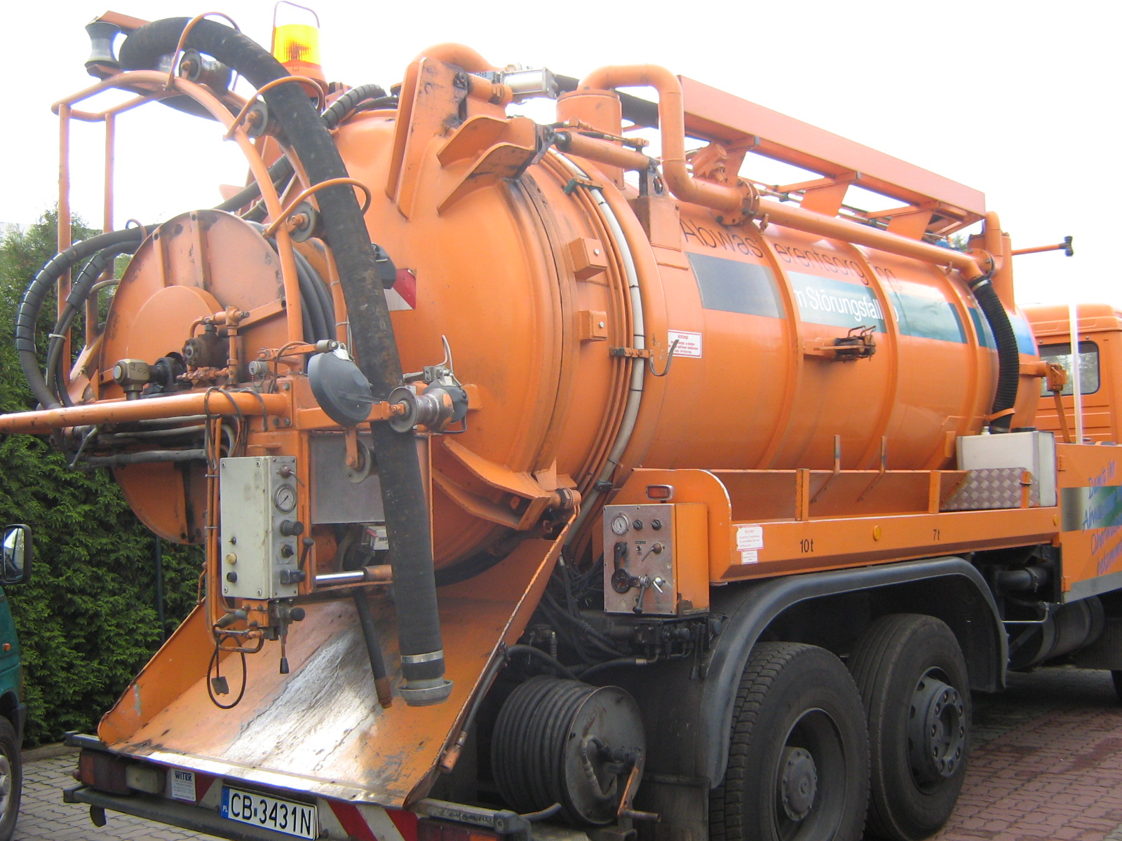 Pomarańczowy samochód asenizacyjny z dużą beczką i grubym wężem, zaparkowany na kostce brukowej, widoczne detale techniczne i oznaczenia pojazdu.