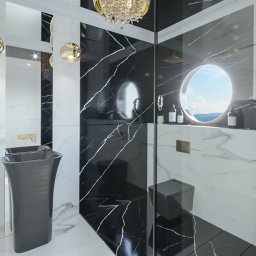 Andrzej Jakubowski - Elegancka łazienka z czarno-białymi marmurowymi płytkami, wolnostojącą umywalką w kolorze grafitowym, złotymi lampami wiszącymi i okrągłym oknem z widokiem na krajobraz.